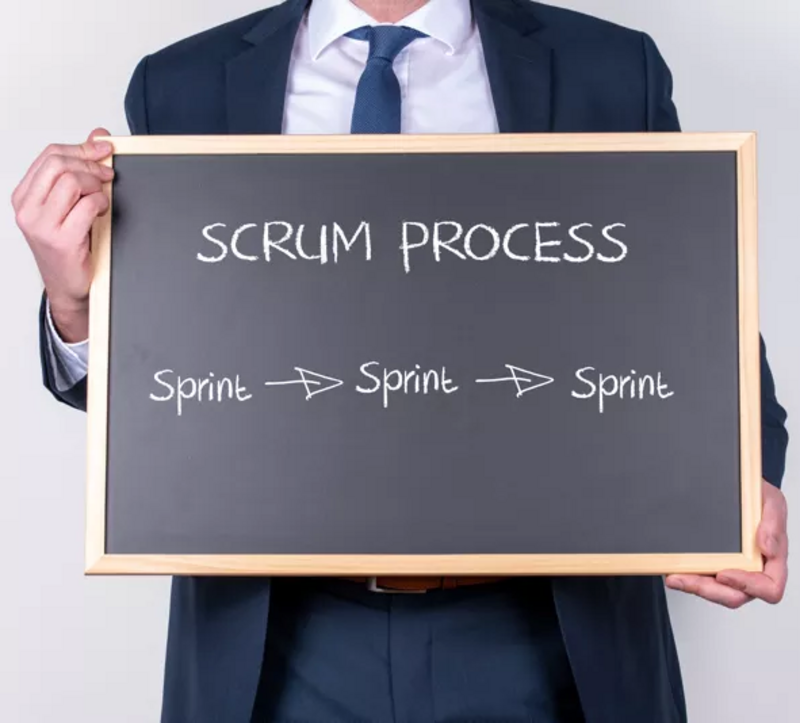 Mann hält Tafel mit seinen Händen. Auf der Tafel steht geschrieben: Srum Process und in einer neuen Zeile: Sprint > Spprint > Sprint 