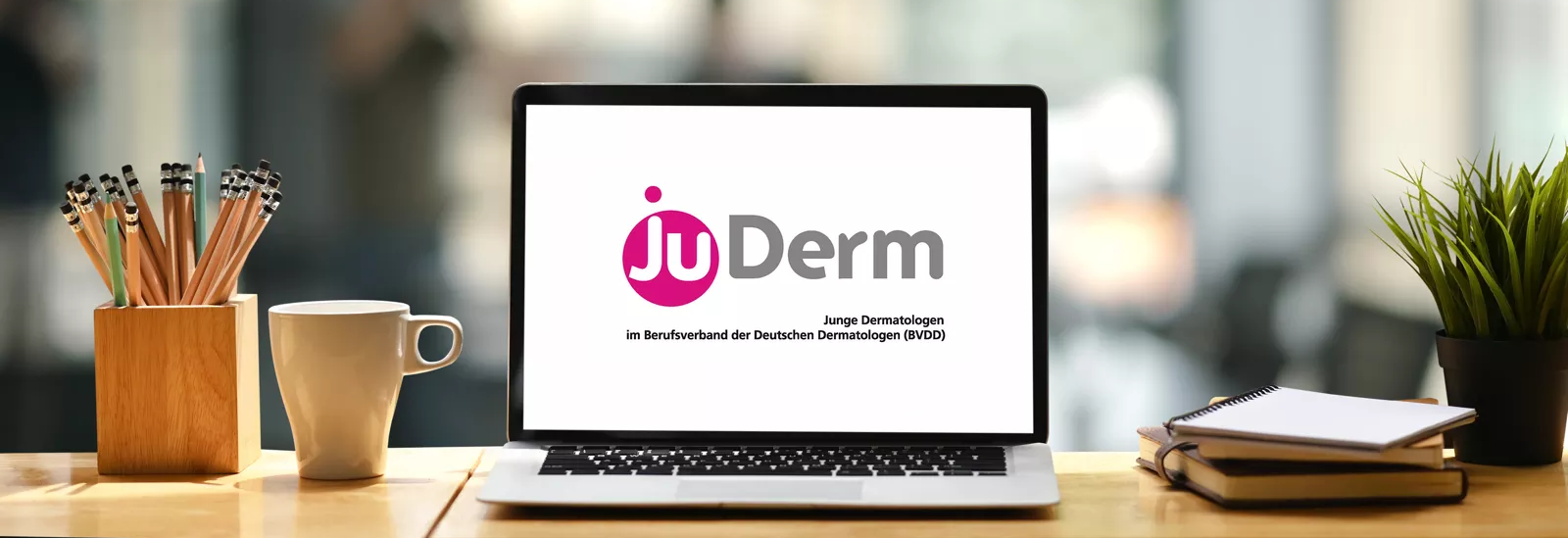 Symbol-Foto Schreibtisch mit Notebook vor unscharfem Hintergrund. Auf dem Display wird das JuDerm Logo angezeigt